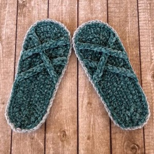Può includere: Un paio di pantofole all'uncinetto color turchese con un design a cinturino incrociato. Le pantofole hanno un aspetto morbido e strutturato e sono rifinite con un bordo grigio chiaro. Le pantofole sono esposte su una superficie di legno.