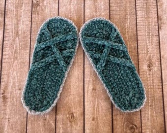 May include: A pair of teal crochet slippers with a crisscross strap design. The slippers have a soft, textured appearance and are trimmed with a light grey border. The slippers are displayed on a wooden surface.