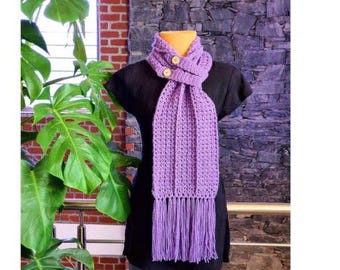 May include: A lavender crochet scarf with fringe and two wooden buttons. The scarf is displayed on a black mannequin. The background includes a brick wall and a purple stone wall. The scarf is a handmade accessory.
