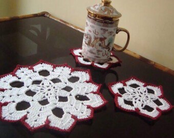 May include: Handmade white and red crochet coasters with a snowflake pattern. A decorative mug with a lid rests on one coaster. These coasters add a touch of elegance to any table setting.
