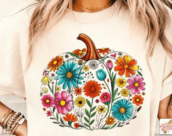 May include: A cream-coloured t-shirt with a colourful floral pumpkin design. The pumpkin is filled with various flowers in shades of blue, orange, pink, and yellow, with a brown stem. A fall-themed graphic tee.