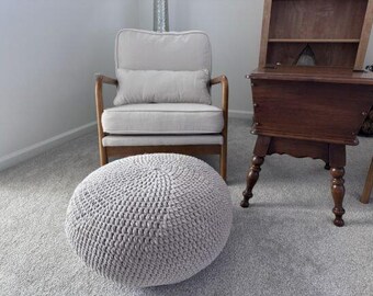 May include: A light grey, round, crocheted ottoman sits on a grey carpet. A light grey armchair with wooden arms and legs is behind the ottoman. A dark wooden side table with a shelf and turned legs is to the right.