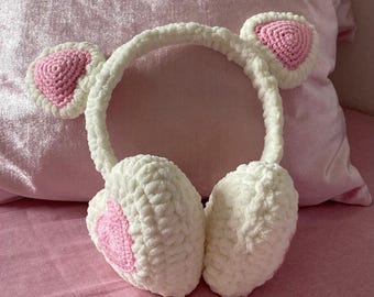 May include: White and pink crochet earmuffs with a headband and ear-shaped accents. The earmuffs feature a heart-shaped design in the center of each ear cover. The background is a pink pillow and pink fabric.