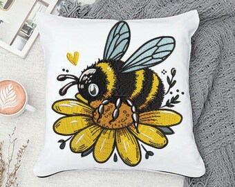 May include: White square cushion featuring an embroidered bee on a yellow flower. The bee has black and yellow stripes and blue wings. A yellow heart is embroidered above the bee. A cup of coffee is visible in the background.