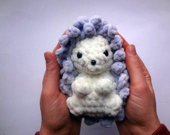 May include: A handcrafted, soft hedgehog toy. The hedgehog features a white body and a light blue, textured exterior. The toy is held in two hands against a white backdrop. The hedgehog has black eyes and a black nose.