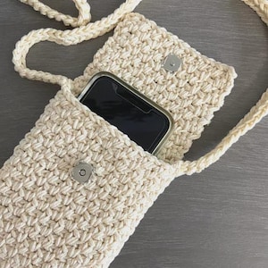 May include: Cream-colored crochet phone crossbody bag with a matching strap. The bag has a flap closure with a silver magnetic snap. A smartphone is visible inside the main compartment. The bag is made of a textured, woven material.