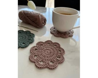 May include: Handmade crochet coasters in mauve and teal. A white mug with a drink rests on a mauve coaster. A ball of yarn and a crochet hook are also visible. The coasters are approximately 10 cm in diameter.