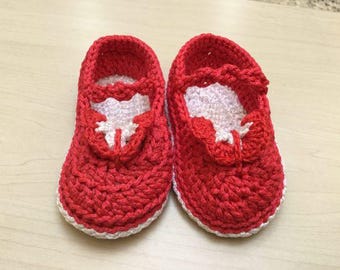 May include: A pair of handmade red crochet baby shoes with white detailing. The shoes have a Mary Jane design with a strap and a decorative element on the front. The soles and edges are trimmed in white.