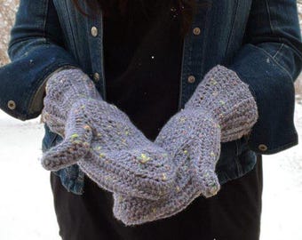 May include: Hand-crocheted mittens in a speckled grey yarn. The mittens have a textured stitch and appear warm. The background shows a denim jacket and a snowy outdoor scene. The mittens are designed to keep hands warm.