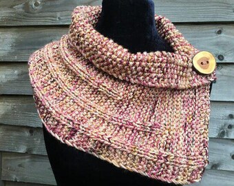 May include: A knitted cowl in pink, beige, and burgundy hues, with a ribbed pattern. It fastens with a large, round wooden button. The neck warmer is displayed on a black mannequin head against a wooden backdrop. A warm accessory.