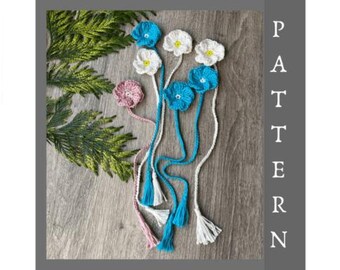 May include: Handmade crochet flower bookmarks in blue, white, and pink with white tassels. The floral bookmarks are displayed on a wooden surface with green foliage. The word "PATTERN" is vertically displayed.