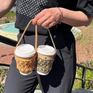 Op de afbeelding: Twee papieren wegwerpbekers voor koffie in een gehaakte houder met twee vakken. De houder heeft een textuurrijke, goudbruine garen hoes aan de linkerkant en een metallic olijfgroene garen hoes aan de rechterkant, beide met bijpassende gevlochten hengsels.