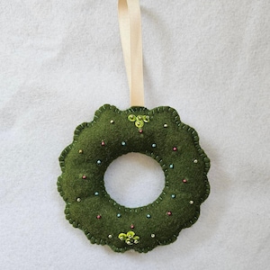 May include: A circular dark green felt wreath ornament featuring a scalloped edge and a light beige ribbon loop for hanging. The surface is decorated with small scattered multi-colored beads and two clusters of bright green sequins at the top and bottom.