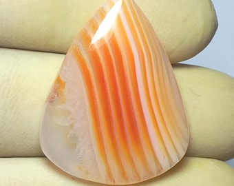 May include: A polished, teardrop-shaped gemstone with alternating bands of orange and white. The stone has a smooth, glossy surface and is held by a gloved hand. The gemstone's pattern resembles a flame or a sunset.