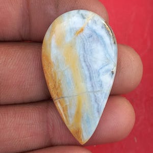May include: A teardrop-shaped gemstone with a polished surface. The stone features a blend of colors, including creamy white, pale blue, and golden yellow, with subtle veining patterns. The gemstone is held in a hand against a red background.