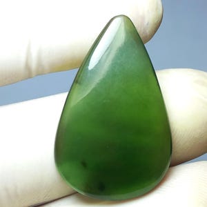 Può includere: Una gemma di giada lucidata a forma di goccia. La pietra è di un verde intenso e traslucido con sottili variazioni di colore. La superficie liscia riflette la luce, evidenziando la sua bellezza naturale. La gemma è tenuta da una mano guantata.