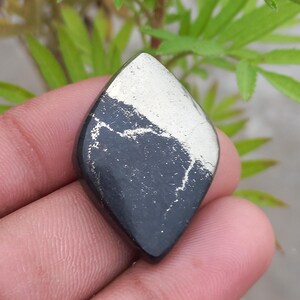 Pode incluir: Uma pedra preciosa polida em forma de losango, com uma base cinza escuro e um topo prateado. A pedra tem uma aparência natural e texturizada, e é segurada em uma mão contra um fundo de folhagem verde.