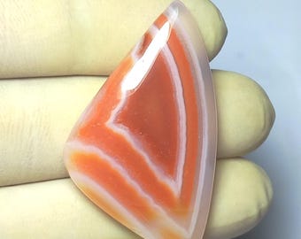 May include: A polished, triangular gemstone with a vibrant orange and white banded pattern. The stone's layers create a chevron design, with a translucent, glossy finish. The gemstone is held in a gloved hand.