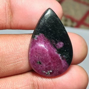 Puede incluir: Una gema en forma de lágrima con una parte superior negra y una base rosa rojiza vibrante. La piedra tiene una superficie pulida, con una mezcla de colores y texturas. La gema se sostiene en una mano.