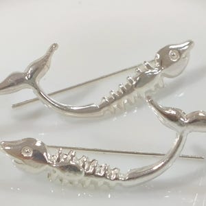 Pode incluir: Dois brincos trepadores prateados em forma de esqueleto de baleia. Os brincos têm um design curvo com uma barbatana caudal numa extremidade e uma cabeça na outra. O detalhe do esqueleto é visível ao longo do corpo curvo.