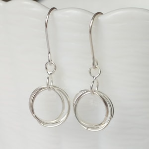 Puede incluir: Un par de pendientes colgantes plateados. Cada pendiente presenta un diseño en bucle con múltiples anillos entrelazados, suspendidos de un simple gancho. Los pendientes se muestran sobre un fondo blanco.