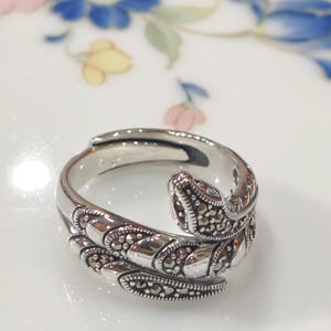 Könnte beinhalten: Ein silberfarbener Ring mit Schlangenmuster. Der Ring umschlingt den Finger und zeigt einen detaillierten Schlangenkopf und -körper. Der Schlangenkörper ist mit kleinen, dunklen Steinen verziert, die Textur und Glanz verleihen. Der Ring ist vor einem weißen Hintergrund platziert.