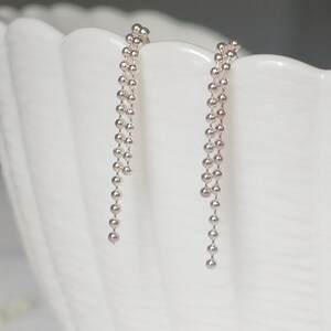 May include: A pair of silver drop earrings. Each earring features a chain of small, round beads that cascade downwards. The earrings are displayed against a white, textured background, creating a contrast that highlights their delicate design.