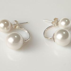 Puede incluir: Un par de pendientes elegantes con dos perlas blancas cremosas y brillantes cada uno. Las perlas están conectadas por un diseño curvo de metal plateado. Los pendientes se muestran sobre una superficie blanca.