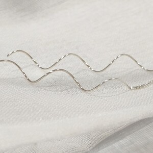 Puede incluir: Un par de delicados pendientes de plata. Cada pendiente presenta un diseño ondulado y retorcido, que se asemeja a una curva suave. Los pendientes están conectados por una pequeña cadena de eslabones plateados. Se muestran sobre una superficie blanca texturizada.