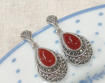 May include: A pair of silver drop earrings. Each earring has a teardrop-shaped, glossy, red stone centre, surrounded by an ornate, filigree design with small, dark accents. The earrings are displayed on a white and blue patterned surface.