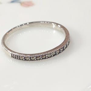 May include: A silver ring featuring a band of small, dark stones. The ring is set against a white surface with a delicate floral pattern. The ring's design is simple and elegant, suitable for everyday wear.