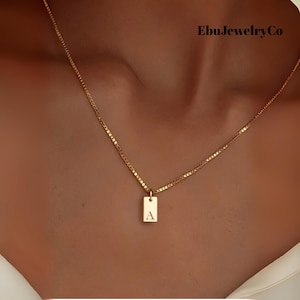 Può includere: Una collana color oro con un ciondolo rettangolare. Il ciondolo ha incisa la lettera "A". La collana è indossata intorno al collo. Il nome del marchio "EbuJewelryCo" è visibile in alto a destra.