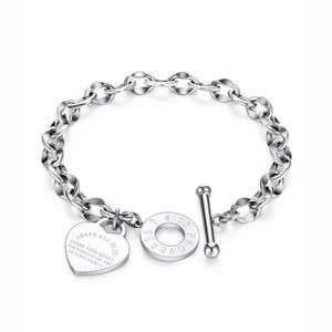 Könnte beinhalten: Ein silberfarbenes Armband mit Knebelverschluss. Das Armband hat einen herzförmigen Anhänger mit dem Text "ABOVE ALL ELSE, GUARD YOUR HEART, FOR EVERYTHING YOU DO FLOWS FROM IT." Ein runder Anhänger liest "SPRÜCHE 4:23".