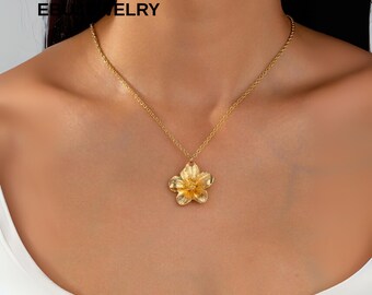 Puede incluir: Collar dorado con un colgante de flor. El colgante es un diseño detallado de flor de cinco pétalos. El collar se lleva puesto, con la cadena y el colgante visibles sobre la piel.