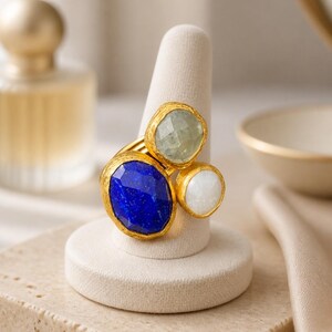 Op de afbeelding: Een goudkleurige ring met drie edelstenen: een grote, ovale, diepblauwe steen, een kleinere, lichtgroene steen en een kleine, witte steen. De ring wordt gepresenteerd op een witte ringhouder, die het ontwerp en het kleurcontrast benadrukt.