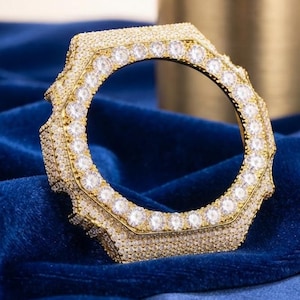 May include: A circular gold-tone watch bezel featuring a faceted, octagonal outer edge. The entire surface is encrusted with numerous small, clear, sparkling stones, with a larger row of round, clear stones lining the inner circular opening.