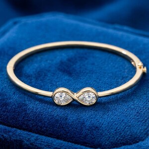 May include: A gold bangle bracelet with an infinity symbol design. The bracelet features two clear, sparkling gemstones set in the center of the infinity symbol. The bracelet is set against a deep blue background.