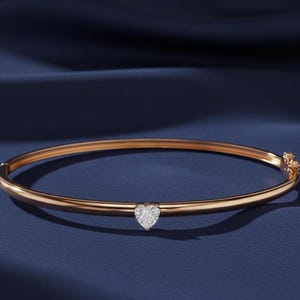 May include: A rose gold bangle bracelet with a heart-shaped diamond pendant. The bracelet is set against a dark blue background, highlighting the jewelry's elegant design. The heart-shaped diamond is the focal point of the bracelet.