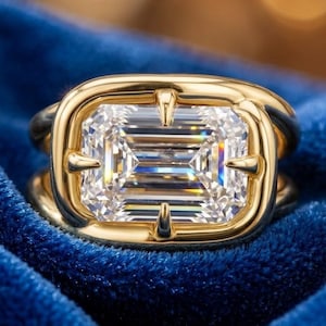 May include: A gold ring featuring a large, rectangular emerald-cut clear gemstone set in a smooth, rounded gold bezel. The setting includes four prominent prongs that secure the stone, creating a bold, polished look with a reflective surface.