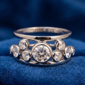 May include: A silver-toned ring featuring a wide band with a double-rail design. The center is set with a large, round, clear faceted stone in a bezel setting, flanked by multiple smaller, round, clear faceted stones in matching bezel settings.