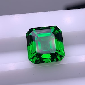 May include: A vivid, emerald-green gemstone with an emerald cut. The gem is square with beveled edges, reflecting light and showcasing its clarity. The gemstone is set against a white surface.