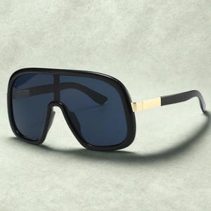 May include: Black oversized sunglasses with dark blue lenses. The sunglasses have a thick black frame with a gold-colored accent on the temples. The lenses are a dark blue color, and the overall design is bold and modern.