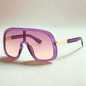 May include: Purple oversized sunglasses with a gradient lens. The frames are a translucent purple with gold-colored accents on the temples. The lenses transition from a light peach to a darker purple.