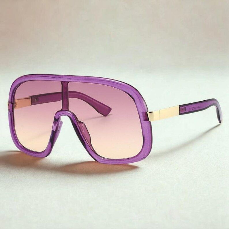 May include: Purple oversized sunglasses with a gradient lens. The frames are a translucent purple with gold-colored accents on the temples. The lenses transition from a light peach to a darker purple.
