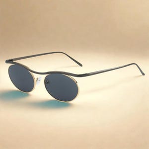 Puede incluir: Gafas de sol con lentes oscuras de forma ovalada enmarcadas en metal dorado. La montura presenta una distintiva barra superior negra ondulada que se conecta a varillas negras delgadas. El puente nasal incluye almohadillas de silicona ajustables.