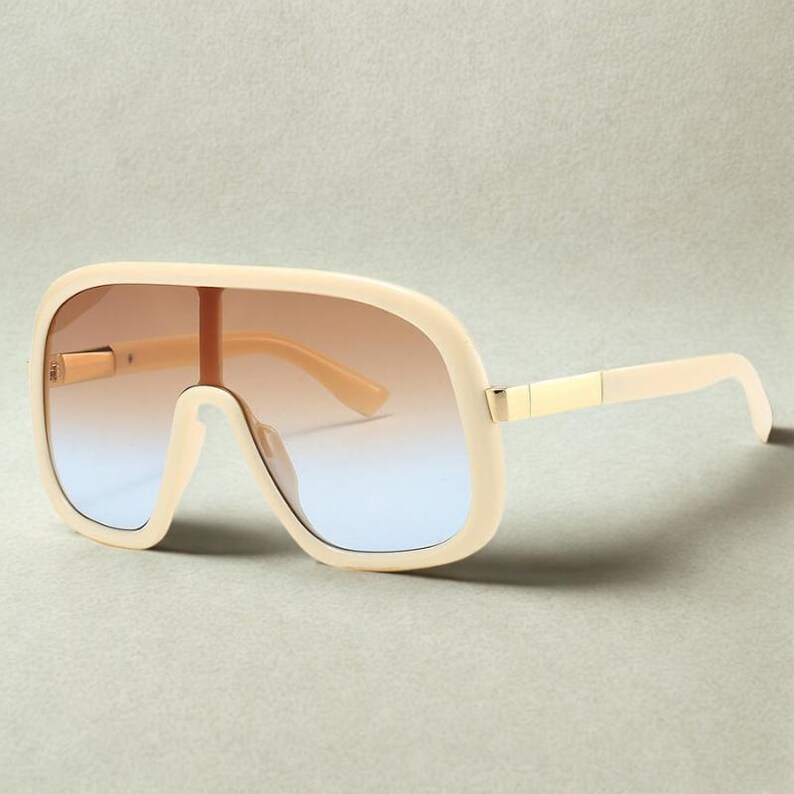 May include: Oversized cream-colored sunglasses with a gradient lens that transitions from brown to blue. The sunglasses have a thick frame and gold-colored metal accents on the hinges. The arms are a matching cream color.