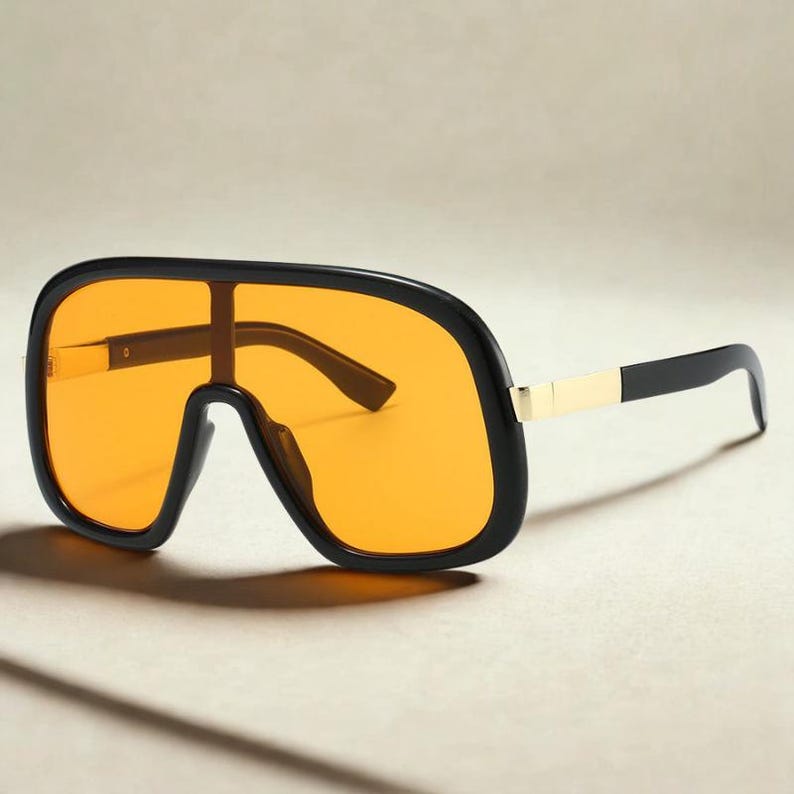 May include: Oversized sunglasses with a black frame and orange lenses. The sunglasses have a thick, rectangular shape with a gold accent on the temples. The arms are black and curve down to the ears.