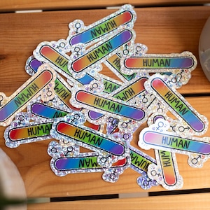 Könnte beinhalten: Eine Ansammlung holografischer Aufkleber mit dem Wort "HUMAN" in schwarzen Großbuchstaben. Die Aufkleber haben einen Regenbogenverlauf und Geschlechtssymbole. Sie liegen auf einer Holzoberfläche.