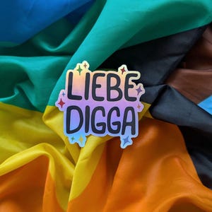 Könnte beinhalten: Ein gestanzter Aufkleber mit dem Text LIEBE DIGGA in fetten schwarzen Buchstaben. Der Aufkleber hat eine holografische, schillernde Oberfläche, die ihre Farbe verändert, und ist mit kleinen, bunten sternförmigen Akzenten verziert.