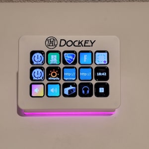 Może przedstawiać: Biały panel sterowania Dockey z wieloma podświetlanymi przyciskami. Na panelu wydrukowano słowo "DOCKEY". Przyciski wyświetlają różne ikony i tekst, w tym "SD OFF", "Studio" i "18:42". Różowe światło emanuje od dołu.